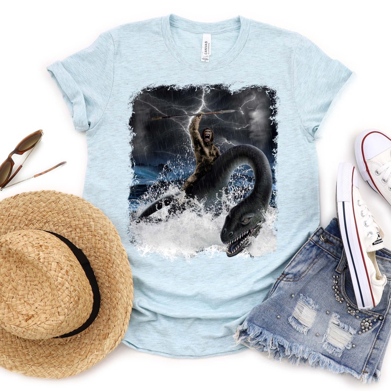 Puede incluir: Una camiseta azul claro con un estampado gr&aacute;fico de un hombre de las cavernas montando un monstruo marino en un mar tormentoso. El gr&aacute;fico est&aacute; desgastado y tiene un aspecto vintage.