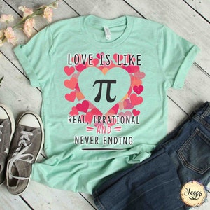 Pu&ograve; includere: Una maglietta verde chiaro con un design a forma di cuore rosa che presenta il simbolo matematico pi. Il testo "Love is like Pi, Real, Irrational and Never Ending" &egrave; stampato sulla maglietta.