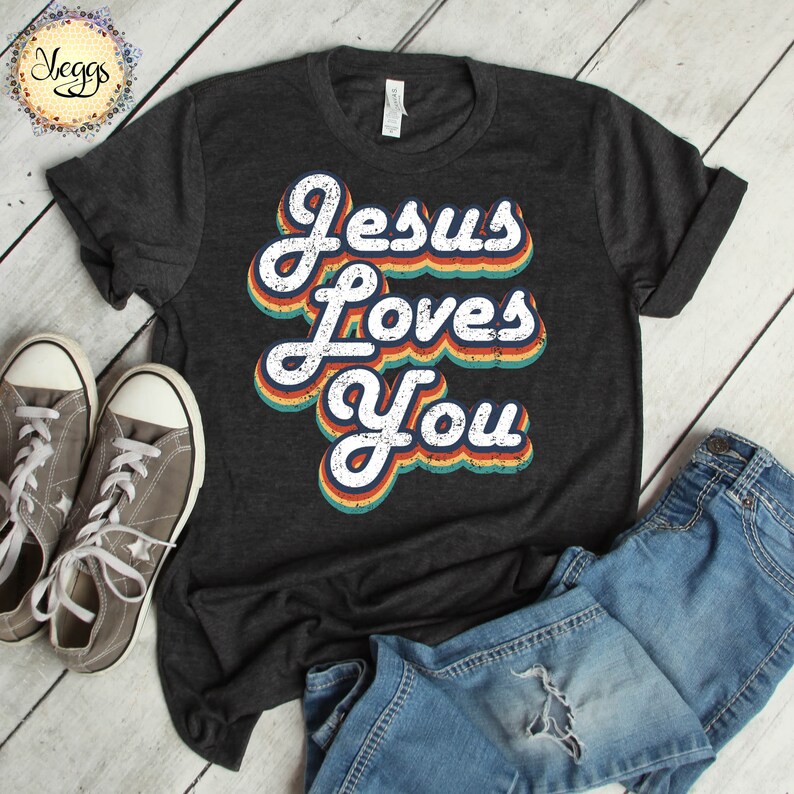Puede incluir: Una camiseta gris oscuro con las palabras "Jesus Loves You" en una fuente retro arco&iacute;ris.