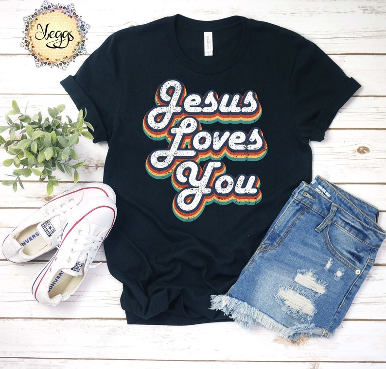 Puede incluir: Una camiseta negra con un gr&aacute;fico de estilo retro que dice "Jesus Loves You" en colores arco&iacute;ris. La camiseta est&aacute; colocada sobre una superficie de madera con un par de zapatillas blancas y pantalones cortos de mezclilla.