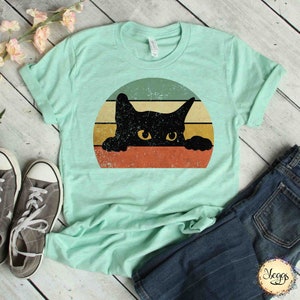Puede incluir: Una camiseta azul turquesa con un gr&aacute;fico de estilo vintage de un gato negro asom&aacute;ndose sobre un fondo rayado naranja, amarillo y verde.
