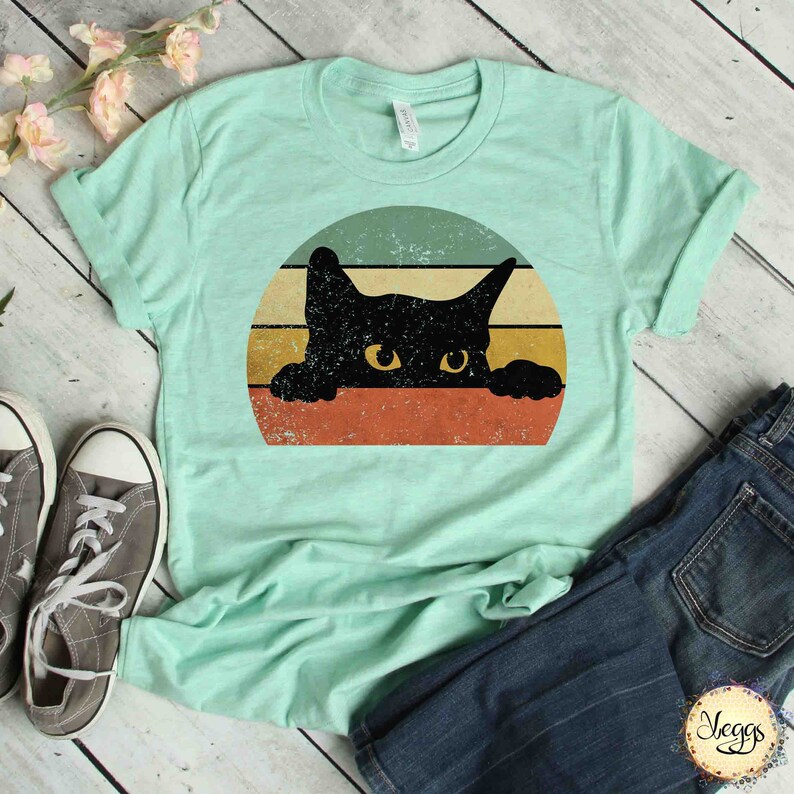 Puede incluir: Una camiseta azul turquesa con un gr&aacute;fico de estilo vintage de un gato negro asom&aacute;ndose sobre un fondo rayado naranja, amarillo y verde.