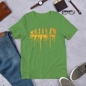 Op de afbeelding: Een groen T-shirt met een gele afbeelding van de evolutie van de mens, eindigend met een persoon die aan een bijenkorf werkt. De afbeelding druipt met gele verf.