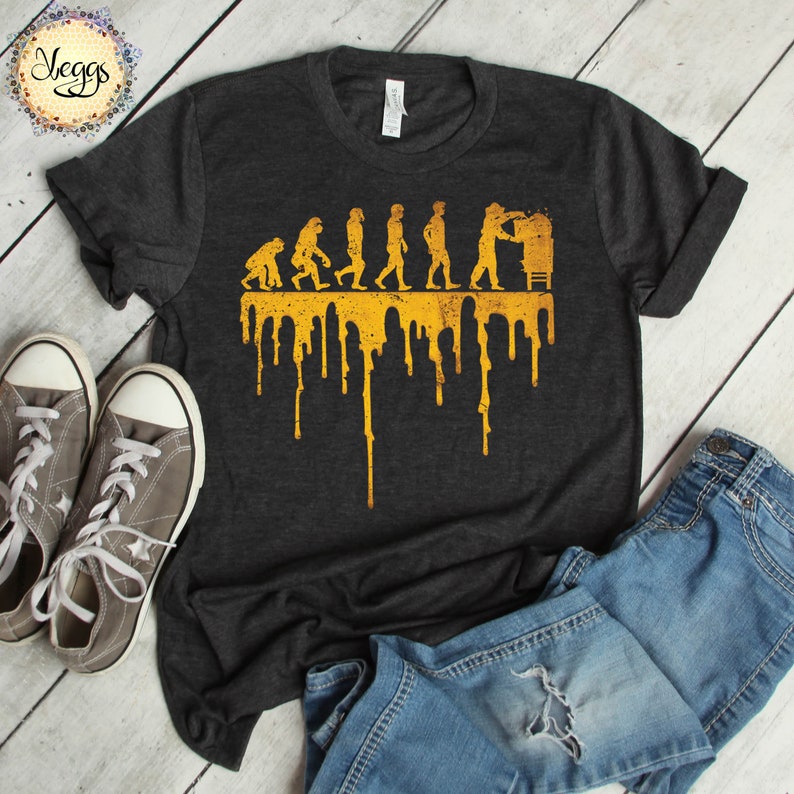 Op de afbeelding: Een donkergrijs T-shirt met een gele afbeelding van de evolutie van de mens, eindigend met een man die een biertje vasthoudt. De afbeelding druipt met gele verf.
