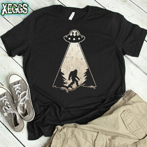 May include: Black t-shirt with a Bigfoot and UFO graphic. The design features a silhouette of Bigfoot in a forest scene, illuminated by a beam from a UFO. The shirt also has the text "XEGGs" in the upper left corner.