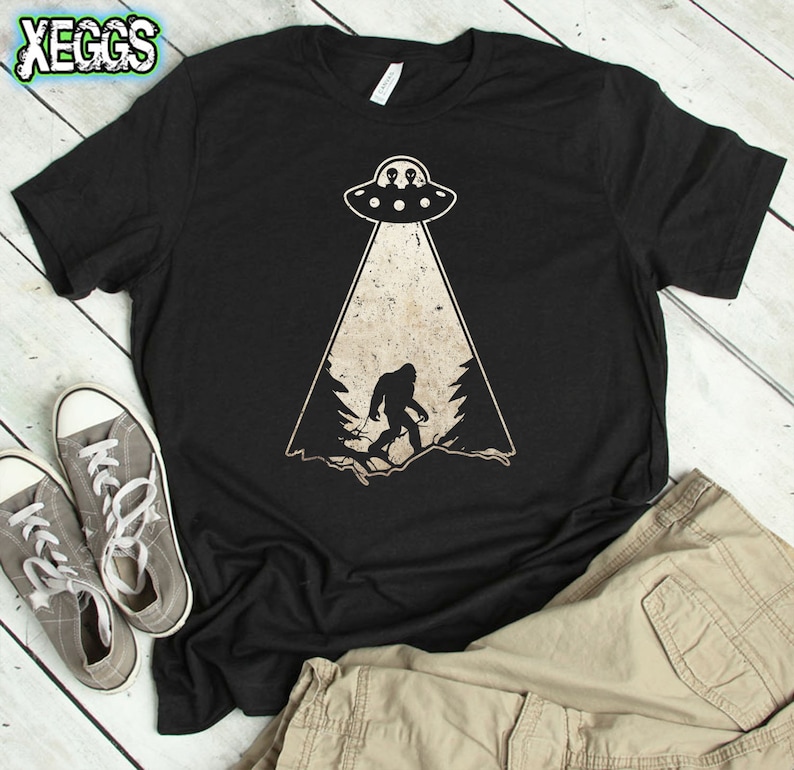 May include: Black t-shirt with a Bigfoot and UFO graphic. The design features a silhouette of Bigfoot in a forest scene, illuminated by a beam from a UFO. The shirt also has the text "XEGGs" in the upper left corner.