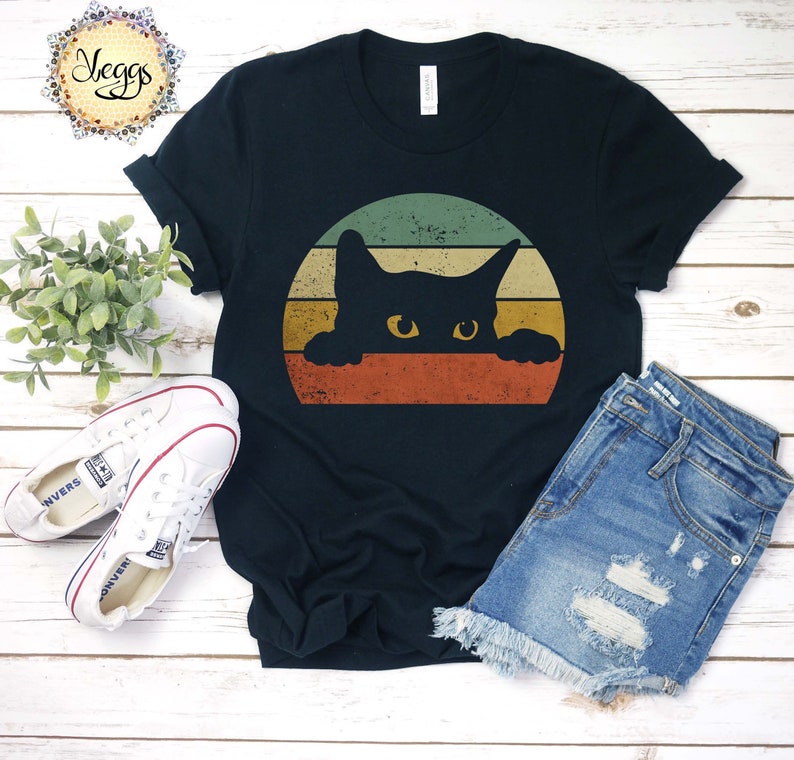 Puede incluir: Una camiseta negra con un gr&aacute;fico de estilo vintage de un gato negro asom&aacute;ndose sobre una puesta de sol. El gato tiene ojos amarillos y mira al espectador.