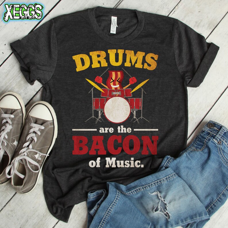 Może przedstawiać: Szary t-shirt z napisem "Drums are the Bacon of Music."  Widoczny jest kresk&oacute;wkowy zestaw perkusyjny z bębnem owiniętym bekonem.