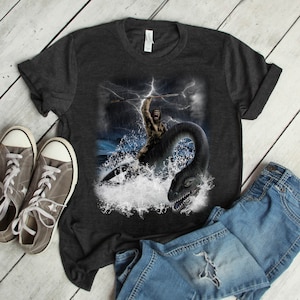 Puede incluir: Una camiseta gris oscuro jaspeada con un estampado gr&aacute;fico de un hombre de las cavernas sosteniendo una lanza y montando un monstruo marino. El monstruo es de color verde oscuro y tiene un cuello y una cola largos. El fondo es un mar tormentoso con rayos.