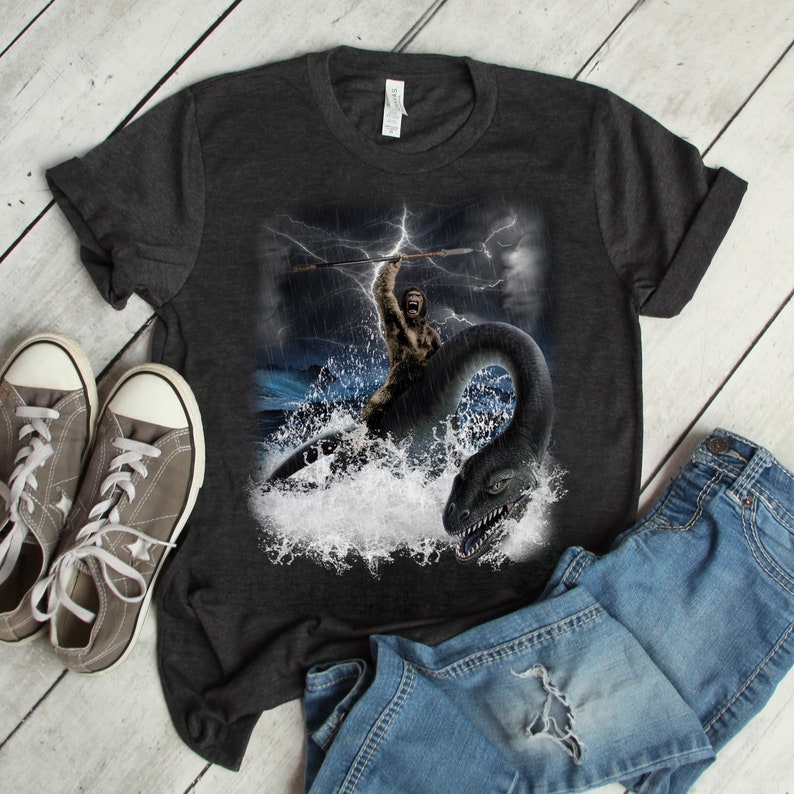 Puede incluir: Una camiseta gris oscuro jaspeada con un estampado gr&aacute;fico de un hombre de las cavernas sosteniendo una lanza y montando un monstruo marino. El monstruo es de color verde oscuro y tiene un cuello y una cola largos. El fondo es un mar tormentoso con rayos.