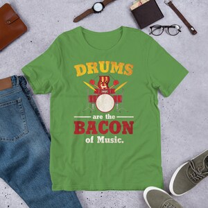 Może przedstawiać: Zielona koszulka z kr&oacute;tkim rękawem z grafiką w stylu vintage zestawu perkusyjnego z tekstem "Drums are the Bacon of Music."