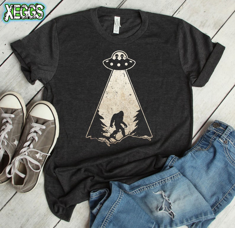 May include: Dark gray t-shirt featuring a Bigfoot graphic being beamed up by a UFO. The design includes a distressed, vintage style with a light beam and forest silhouette. The shirt is paired with gray sneakers and blue jeans.