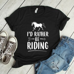 Puede incluir: Camiseta negra con la frase "I'd Rather Be Riding" y un dibujo de un caballo blanco. La imagen incluye zapatillas grises y vaqueros azules, creando una est&eacute;tica casual de tem&aacute;tica ecuestre.