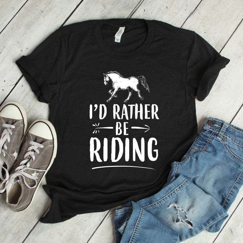 Puede incluir: Camiseta negra con la frase "I'd Rather Be Riding" y un dibujo de un caballo blanco. La imagen incluye zapatillas grises y vaqueros azules, creando una est&eacute;tica casual de tem&aacute;tica ecuestre.