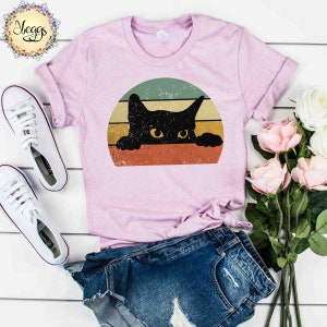 Puede incluir: Una camiseta rosa con un gato negro asom&aacute;ndose por encima de un gr&aacute;fico de puesta de sol de estilo vintage. El gato tiene ojos amarillos y mira directamente al espectador.