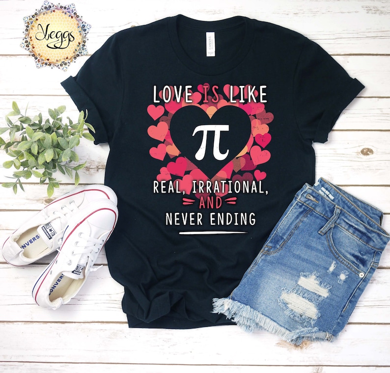 Pu&ograve; includere: Maglietta nera con un design a forma di cuore rosa che presenta il simbolo matematico pi. Il testo "LOVE IS LIKE" &egrave; sopra il cuore e il testo "REAL, IRRATIONAL, =AND= NEVER ENDING" &egrave; sotto il cuore.