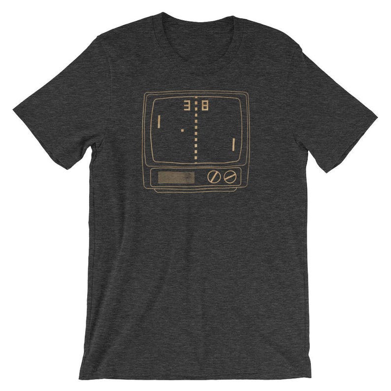 Puede incluir: Una camiseta gris oscuro con una ilustraci&oacute;n de estilo vintage de un juego de arcade retro. El juego se muestra en una pantalla de televisi&oacute;n con el marcador 3:8.