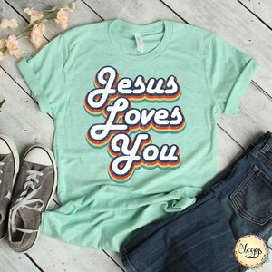 Puede incluir: Una camiseta verde claro con un gr&aacute;fico de estilo retro que dice "Jes&uacute;s te ama" en colores arco&iacute;ris.
