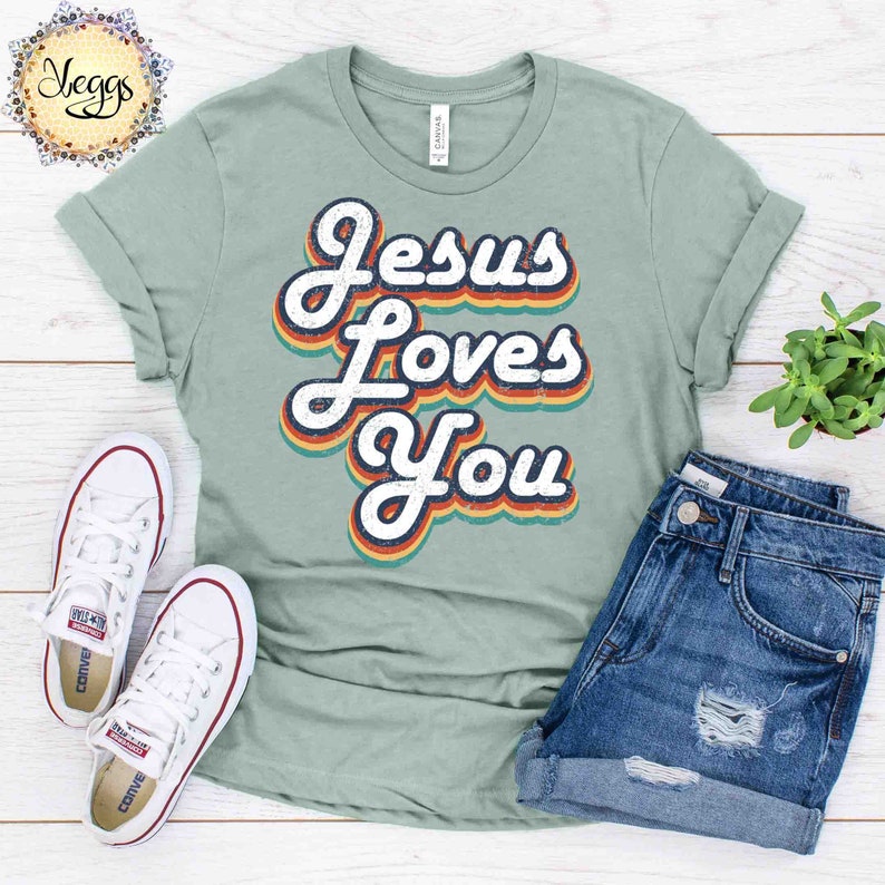 Puede incluir: Una camiseta azul claro con el texto "Jesus Loves You" en una fuente retro arco&iacute;ris. El texto tiene un aspecto desgastado y est&aacute; centrado en la camiseta.