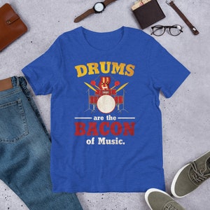 Może przedstawiać: Koszulka w kolorze kr&oacute;lewskiego błękitu z grafiką w stylu vintage, na kt&oacute;rej widnieje napis "Drums are the Bacon of Music." Grafika przedstawia kresk&oacute;wkowy zestaw perkusyjny z kawałkiem bekonu grającym na perkusji.