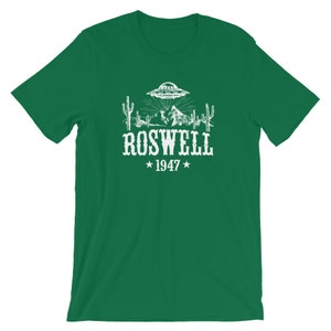 May include: Green t-shirt with a white graphic of a UFO over a desert landscape with cacti and mountains. The text "ROSWELL *1947*" is below the graphic.