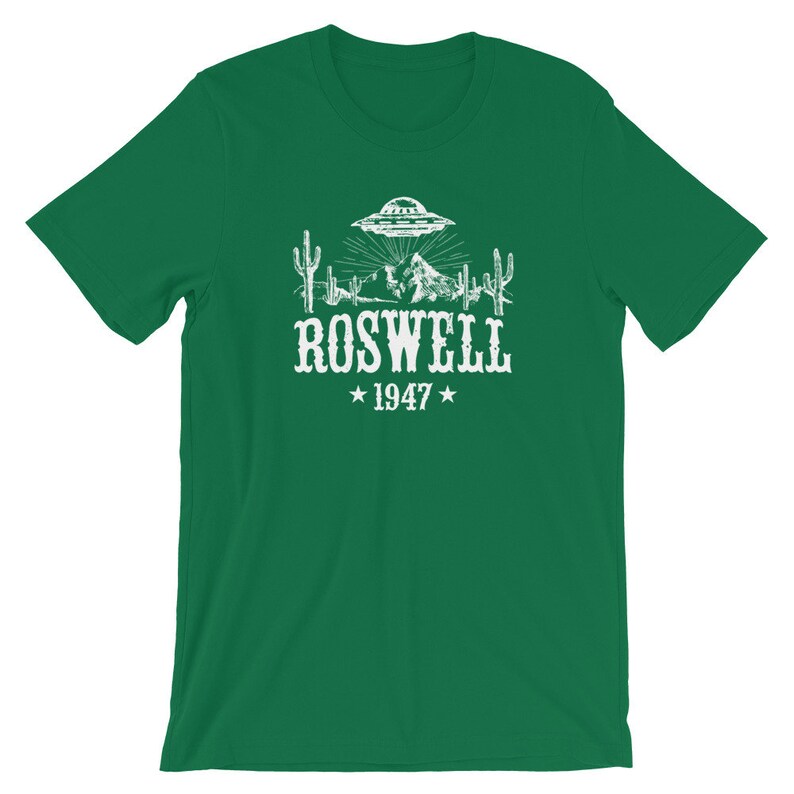 May include: Green t-shirt with a white graphic of a UFO over a desert landscape with cacti and mountains. The text "ROSWELL *1947*" is below the graphic.