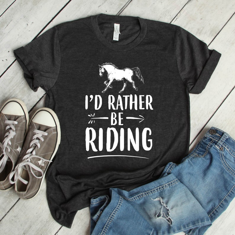 Puede incluir: Camiseta gris oscuro con la frase "I'd Rather Be Riding" y un dibujo de un caballo blanco. La imagen incluye zapatillas grises y vaqueros azules, sugiriendo un estilo casual de tem&aacute;tica ecuestre.