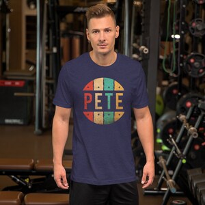 May include: A navy blue t-shirt with a vintage-style circle graphic featuring the name "PETE" in white letters. The circle has a red, orange, yellow, green, and blue color scheme with white stars around the edge.
