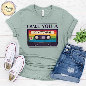 May include: A light green t-shirt with the text "I made you a mixtape" and a retro cassette tape graphic. The cassette tape is black with rainbow colored stripes and a white label with the letter "A" on it.
