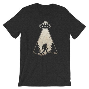 May include: Dark gray t-shirt featuring a Bigfoot graphic being abducted by a UFO. The design includes a UFO with two aliens, a spotlight, and a silhouette of Bigfoot in a forest setting. The shirt is made of a soft material.