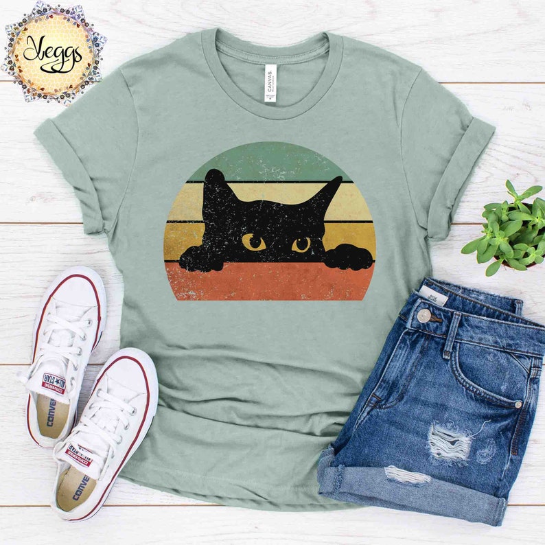 Puede incluir: Una camiseta verde claro con un gr&aacute;fico retro de un gato negro asom&aacute;ndose por encima de una puesta de sol colorida. El gato tiene ojos amarillos y mira directamente al espectador.