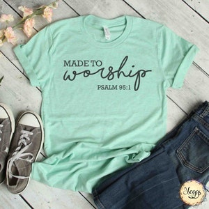 May include: A light green t-shirt with the text "MADE TO worship PSALM 95:1" in black lettering. The shirt is laid out on a wooden surface with a pair of gray sneakers and blue jeans.