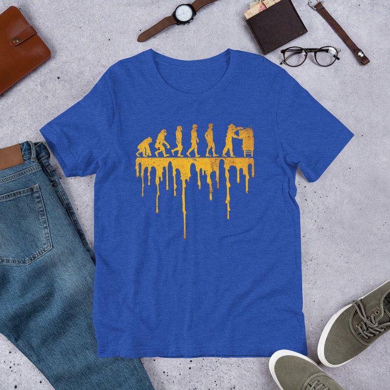 Op de afbeelding: Een koninklijk blauw t-shirt met een gouden afbeelding van de evolutie van de mens, eindigend met een persoon die aan een computer werkt. De afbeelding druipt met gouden verf.
