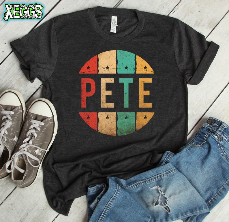 May include: A dark grey t-shirt with a vintage-style circular design featuring the name "PETE" in bold, red, yellow, green, and blue lettering. The circle is surrounded by small white stars.