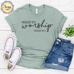 May include: A light green t-shirt with the text "MADE TO worship PSALM 95:1" in black lettering. The shirt is laid out on a wooden surface with a pair of white sneakers and denim shorts.