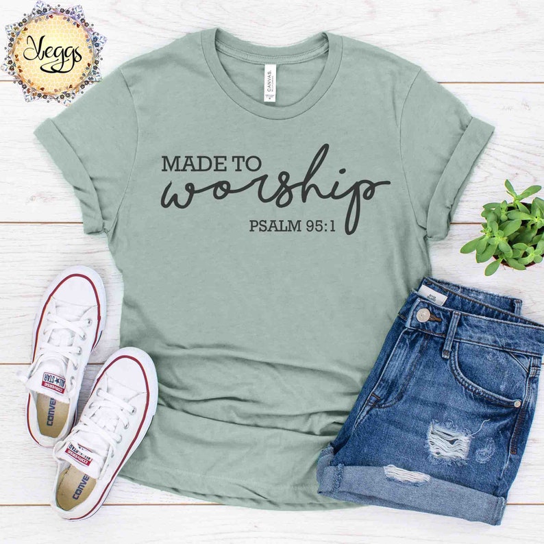 May include: A light green t-shirt with the text "MADE TO worship PSALM 95:1" in black lettering. The shirt is laid out on a wooden surface with a pair of white sneakers and denim shorts.