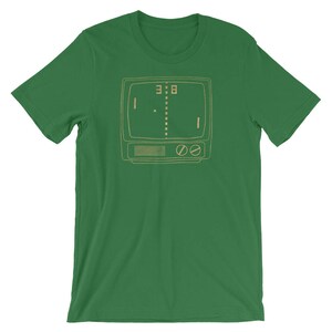 Puede incluir: Una camiseta verde con una ilustraci&oacute;n de estilo retro de una pantalla de televisi&oacute;n vintage que muestra un videojuego simple. El juego muestra dos paletas y una pelota en el medio de la pantalla. El marcador es 3-8.