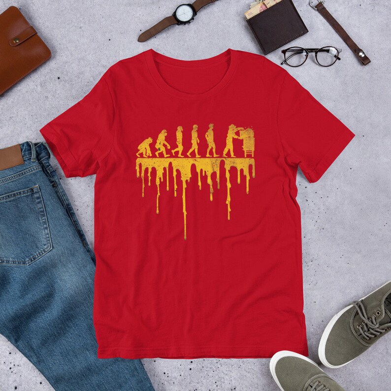 Op de afbeelding: Rood T-shirt met een gele afbeelding van de evolutie van de mens, eindigend met een persoon die aan een computer werkt. De afbeelding heeft een druppelend effect.