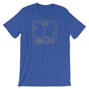 Puede incluir: Una camiseta azul real con un contorno de estilo vintage de una pantalla de televisi&oacute;n que muestra un simple juego de pong pixelado. El marcador es de 3 a 8.