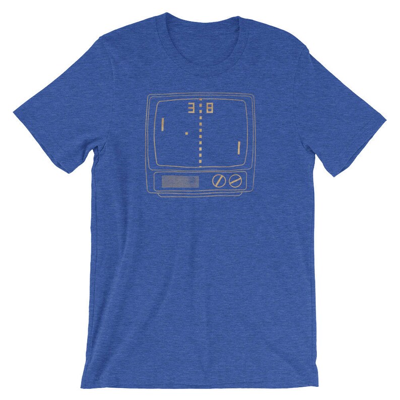 Puede incluir: Una camiseta azul real con un contorno de estilo vintage de una pantalla de televisi&oacute;n que muestra un simple juego de pong pixelado. El marcador es de 3 a 8.