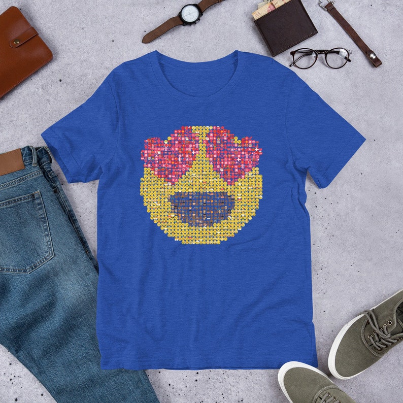 Puede incluir: Camiseta azul real con una cara de emoji grande. El emoji tiene ojos en forma de coraz&oacute;n y est&aacute; formado por caras de emoji m&aacute;s peque&ntilde;as en tonos amarillos, rosas y morados. La camiseta se muestra sobre una superficie gris con otros accesorios.