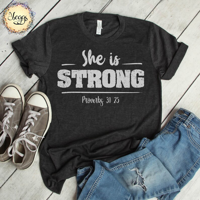 May include: A dark heather grey t-shirt with a white graphic that says "She is STRONG" with "Proverbs 31:25" underneath. The shirt is laid out on a wood surface with a pair of grey sneakers and blue jeans.
