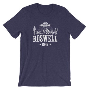 May include: A navy blue t-shirt with a white graphic of a UFO over a desert landscape with cacti and mountains. The text "ROSWELL 1947" is below the graphic.