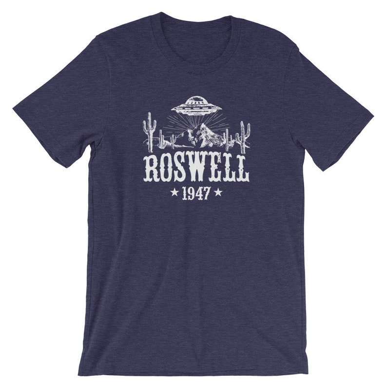 May include: A navy blue t-shirt with a white graphic of a UFO over a desert landscape with cacti and mountains. The text "ROSWELL 1947" is below the graphic.