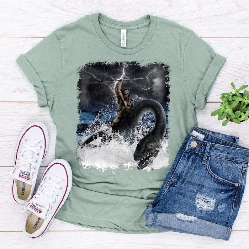 Puede incluir: Una camiseta verde claro con un estampado gr&aacute;fico de un hombre de las cavernas sosteniendo una lanza y montando un monstruo marino en un mar tormentoso.