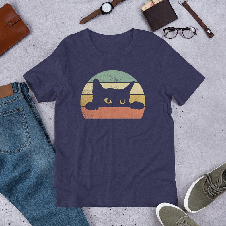 Puede incluir: Una camiseta azul marino con un gr&aacute;fico de estilo vintage de un gato negro asom&aacute;ndose por encima de una puesta de sol. El gato tiene ojos amarillos y est&aacute; silueteado contra una puesta de sol multicolor.
