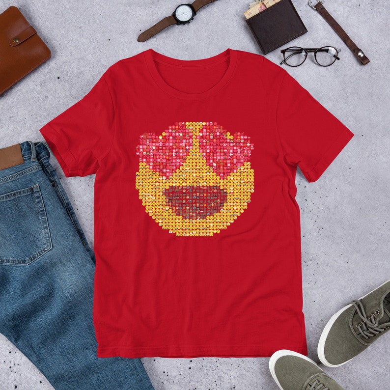 Puede incluir: Camiseta roja con un gran dise&ntilde;o de emoji. El emoji tiene ojos en forma de coraz&oacute;n y una boca sonriente, creada a partir de peque&ntilde;os cuadrados. La camiseta se muestra sobre una superficie gris con otros accesorios.