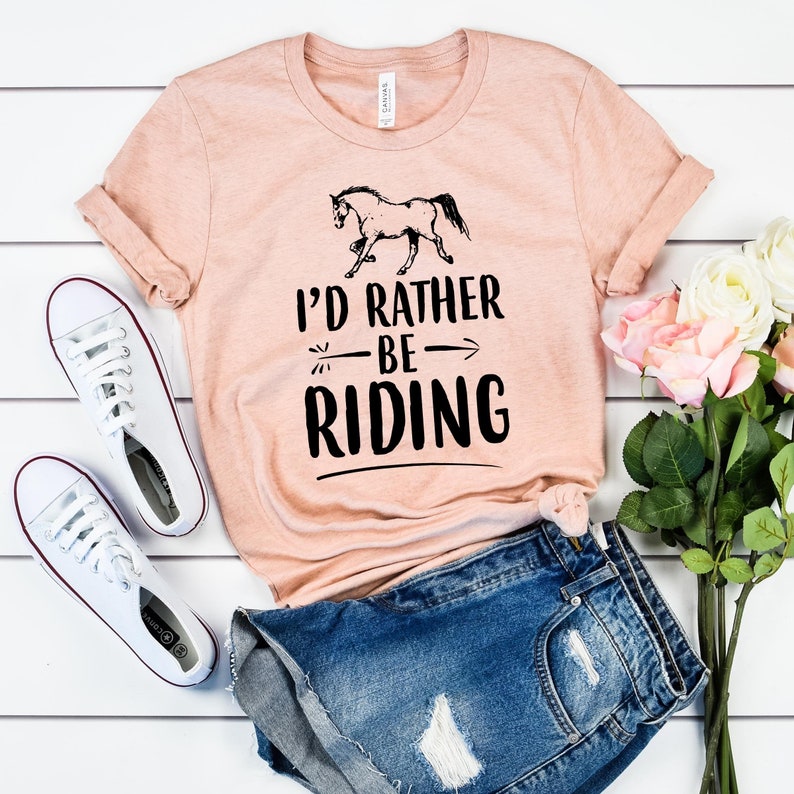 Puede incluir: Una camiseta color melocot&oacute;n con el texto "I'D RATHER BE RIDING" y un gr&aacute;fico de un caballo negro. La camiseta se combina con pantalones cortos de mezclilla y zapatillas blancas. Tambi&eacute;n hay un ramo de flores rosas y blancas.