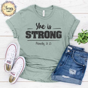 May include: A light green t-shirt with the text "She is STRONG" in black with a distressed look. Below the text is "Proverbs 31:25" in black.