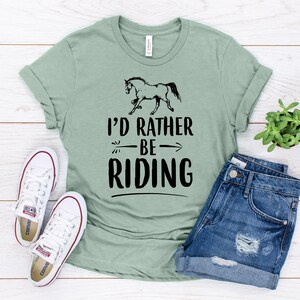 Puede incluir: Camiseta verde salvia con la frase "I'd Rather Be Riding" y un gr&aacute;fico de un caballo negro. La imagen incluye pantalones cortos de mezclilla y zapatillas de lona blancas, creando una est&eacute;tica ecuestre relajada.